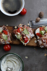 Three toasts with bacon, cherry tomatoes, cucumber and cream cheese. Sandwiches on a wooden board on a dark table. Snacks, top view.