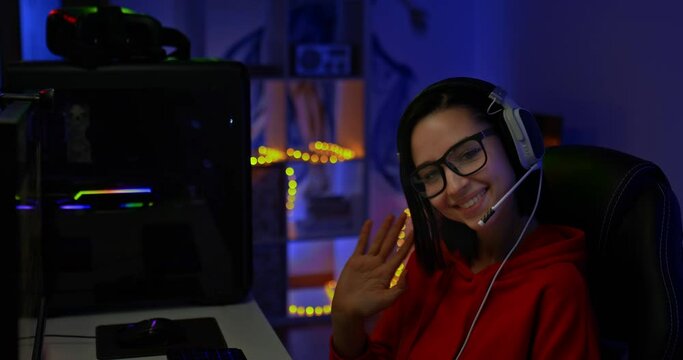 Portrait, a beautiful girl blogger waves her hand and smiles, greets the participants of the online broadcast. Girl with glasses and headphones sits in a dark room with illumination, virtual