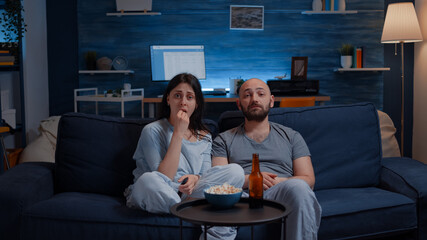 Amazed couple watching movie on TV at night and eating popcorn, drinking beer discussing on film...