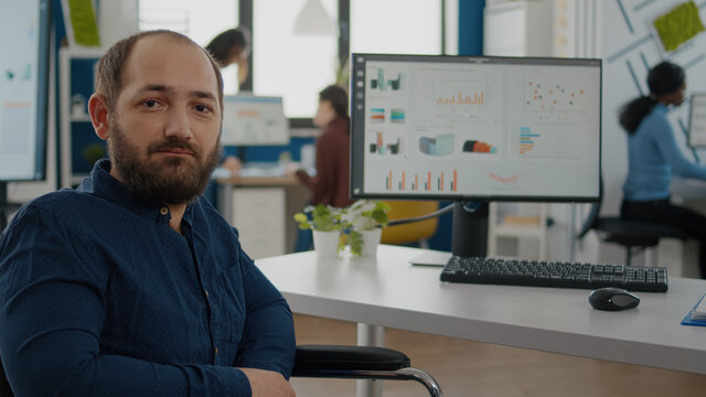 Paralized, invalid, handicapped office manager looking at camera sitting in modern startup company office in wheelchair working with team at financial project. Tired disabled immobilized businessman