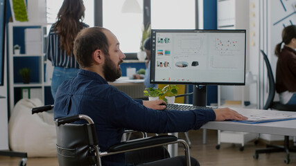 Immobilized paralysed handicapped manager analysing graphs and typing on computer sitting in wheelchair in start up business office, while diverse team working at financial project in background.