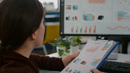 Business woman with disabilities working for deadline project reading documents from clipboard checking graphs sitting immobilized paralized in wheelchair in business office looking on computer