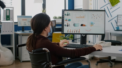 Disabled handicapped businesswoman with face mask immobilized in wheelchair working at financial project typing on pc showing data, prosessing on display sitting in new normal startup business office.