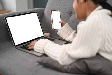 Cropped shot of cheerful young female holding mobile phone and using laptop computer on couch.