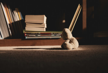 Teddy bear is sitting down on carpet next to blurry bookshelf background in retro filter,Lonely teddy bear stay home alone in living room at night, lonely concept, international missing children's day
