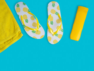 White flip-flop sandals with pineapple decor, bright yellow bottle of sunscreen lotion and beach towel on vivid sky blue background. Summer season vacation concept. Image with copy space, horizontal