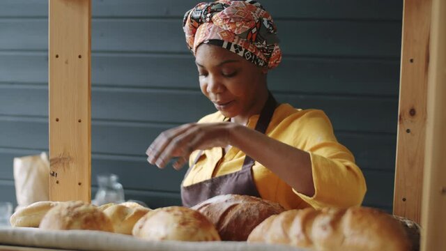 Young beautiful Afro-American woman putting loafs of freshly baked bread on shelve and smiling while working in bakery
