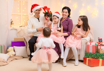 family portrait of a parents and children, dressed in santa helper hat, sitting on a couch in home interior decorated with christmas lights and new year holiday gifts