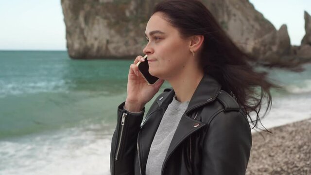Angry young brunette woman gets angry, screams and swears while talking emotionally on the phone. Seascape, stormy sea background