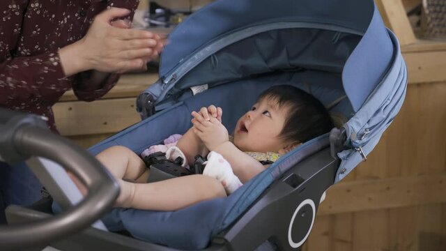 cropped shot innocent Korean baby lying in the stroller at home is looking at her mom who is interacting with her by singing and clapping hands.