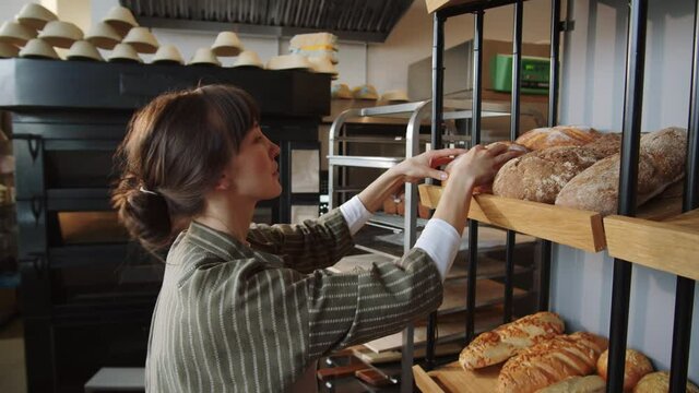 Young caucasian woman in apron putting freshly baked loafs of bread on shelves of display rack while working in bakery