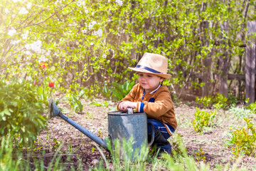 Cute little toddler boy in a hat and rubber boots is watering plants with a watering can in the garden. A charming little kid helping his parents grow vegetables.