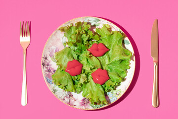 Green coral lettuce  leaves with bright red lips figurines on plate and golden fork and knife on bold pink background. Creative Valentines day,summer or spring romantic idea with kiss.