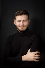 Studio portrait of blue-eyed smiling young handsome man in black clothes on black background; portrait to the waist with hands; a guy sitting on a chair; look directly into the camera lens