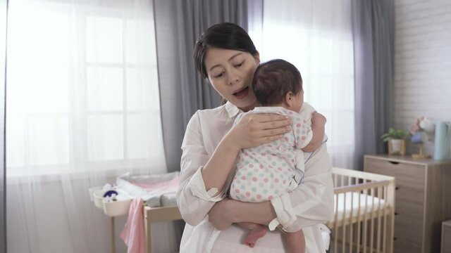 Korean mom is holding and coaxing her fussy newborn daughter while burping with patience at background bright baby bedroom interior.