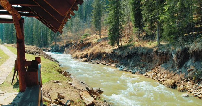 On the left, on the low bank of the mountain river, there is a gazebo for relaxing. The right bank of the river is steeper and covered with forest. The river originates in the mountains.