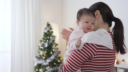portrait adorable baby is burying her face in mommy’s shoulder while being patted on the back for burping in cozy home interior on christmas morning.