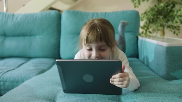 Preschooler Girl Playing Games Using Digital Tablet Lying On Sofa Alone. Curious Little Kid Girl Hold Pad Computer Watching Video At Home Children Education Or Tech Internet Addiction Concept
