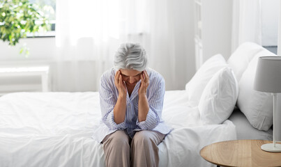 health, old age and people concept - senior woman in pajamas suffering from headache sitting on bed...