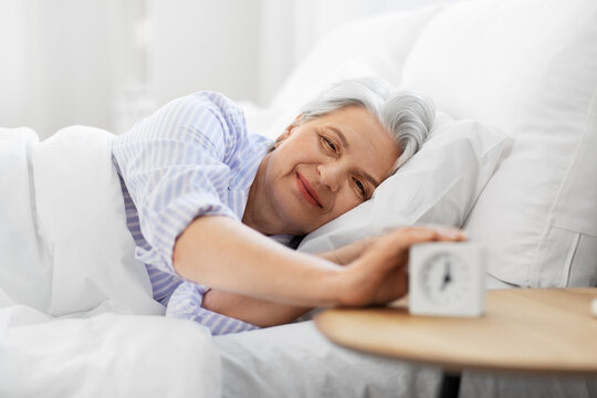 old age and people concept - happy smiling senior woman with alarm clock lying in bed at home bedroom