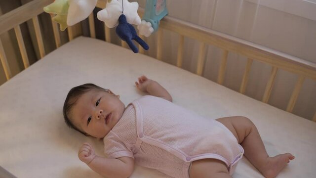 cropped adorable female infant in bodysuit is lying alone in the baby cot and looking at the colorful mobiles in the bedchamber at home.