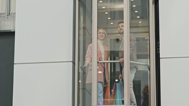 Tracking full-shot of stylish young Caucasian couple with lots of shopping bags going down glass elevator in modern shopping centre