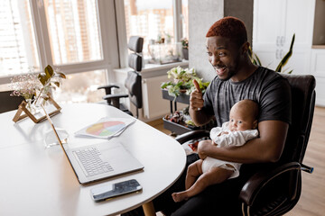 Excited male employee is sitting at the desk with baby son at home, using computer, having a video conference. Happy young businessman talking with colleagues at online meeting, discussing a project