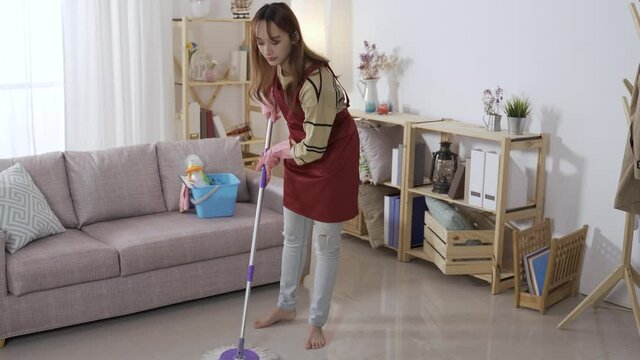 full length Chinese female in an apron is standing and mopping the floor with a swabber in a bright home interior at daytime.