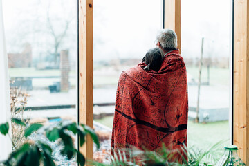 back view of a senior couple at home wrapped in a blanket while looking outside from a big window