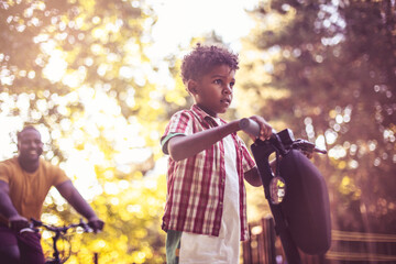 African dad riding bike and his son riding e-scooter. Focus is on little boy.