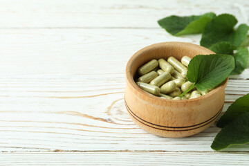 Bowl with herbal capsules and leaves on white wooden background