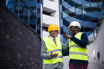  Mature engineer discussing the structure of the building with architects colleague at construction site. Businessmen talking.