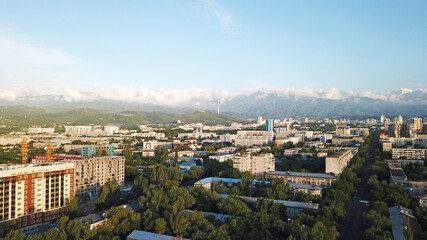 Fototapeta premium Bright color sunset over the city of Almaty. Huge clouds over the mountains and the city shimmer from bright blue to yellow and dark blue. Tall houses and green trees, cars driving on the roads.
