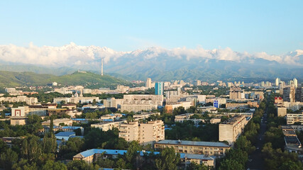 Fototapeta na wymiar Bright color sunset over the city of Almaty. Huge clouds over the mountains and the city shimmer from bright blue to yellow and dark blue. Tall houses and green trees, cars driving on the roads.