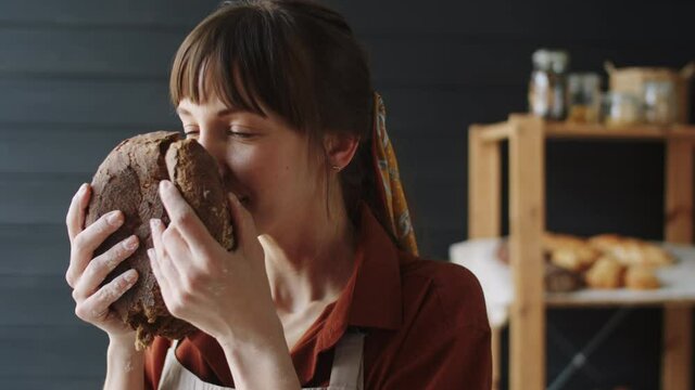 Zoom in shot of young happy female baker in apron holding round loaf of freshly baked rye bread and enjoying its smell