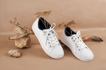 Ethical vegan shoes concept. A pair of white sneakers with dry flowers on the wood and pile of stones, neutral beige craft paper background - obrazy, fototapety, plakaty