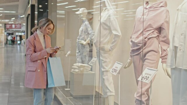 Medium shot of young pretty woman wearing smart casual clothes standing at window display of fashionable clothing store taking photo of mannequin on smartphone