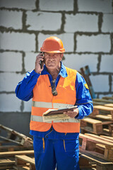 Male engineer reviewing blueprints on clipboard at construction site using mobile phone