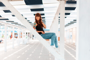 Young beautiful woman with tattoos on her arm, posing, sitting on a white staircase. In the background is an architectural perspective. Outdoor
