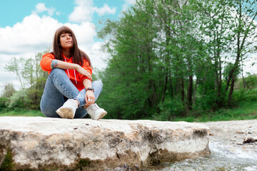 A young Caucasian woman sits on a rock on the river bank. Bottom view. Park and outdoor activities