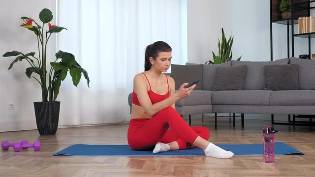 Sportive beautiful woman in sportswear sitting on yoga blue mat uses smartphone. Fit girl athlete resting relax after active intense sport fitness workout exercise training at home in living room