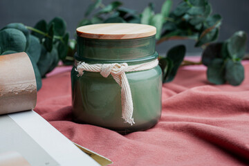 Scented candle in a glass jar on a background of kraft paper, green leaves and a pine cone. Light text background