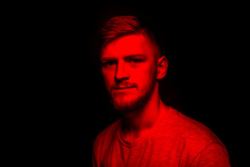 Young attractive male person posing in contrast red light. Portrait of handsome guy in dark studio with red neon lightning. Confident pretty man looks at camera, fashion beauty art concept