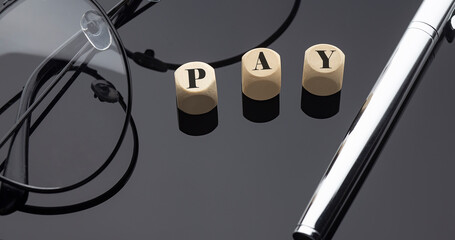 PAY concept, words on wooden blocks on the black background with pen and glasses.