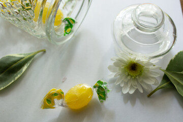 Still life with a glass vessel, white flowers, leaves and yellow sweets on a white table