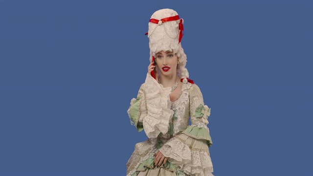 Portrait of a courtier lady in a white vintage lace dress and wig talking on her smartphone. Young woman posing in studio with blue screen background. Close up. Slow motion ready 59.94fps.
