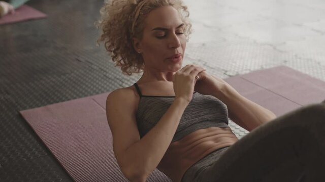 Medium long of blonde curly Caucasian female athlete wearing sports bra, lying on yoga mat on rubber gym flooring, doing sit-ups