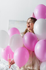 Fototapeta na wymiar Excited woman with champagne looking away near festive balloons in bedroom
