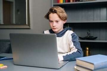 Portrait of cute pupil boy using typing laptop computer doing online lesson via Internet at desk, selective focus. Child schoolboy doing homework at home. Concept of remote distance education.