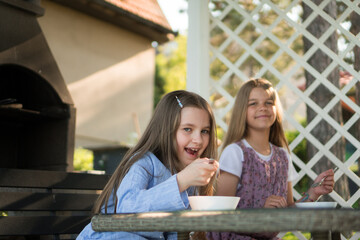 Two adorable little girls are sitting at the table in the backyard and eating soup.